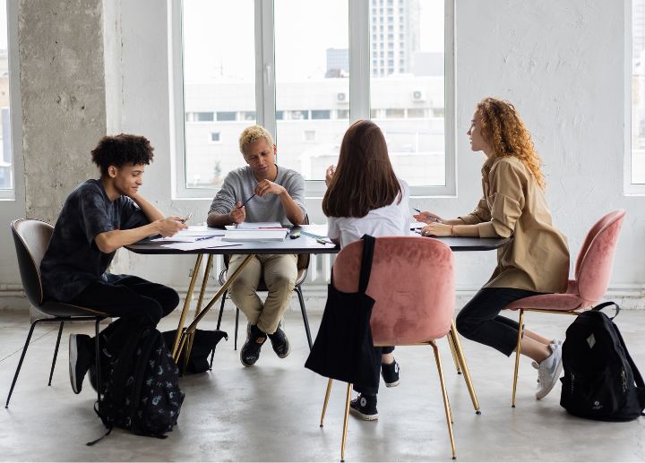 Group of people sitting around a table in a bright room, working together with papers and laptops.